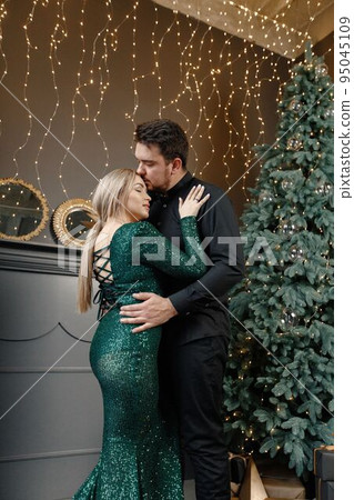 Young woman and man standing near christmas tree and hugging. Cheerful family celebrating Christmas. Blonde woman wearing fashion green dress and bearded man wearing black shirt. 95045109