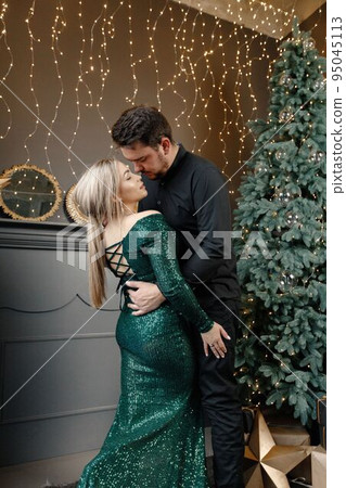 Young woman and man standing near christmas tree and kissing. Cheerful family celebrating Christmas. Blonde woman wearing fashion green dress and bearded man wearing black shirt. 95045113