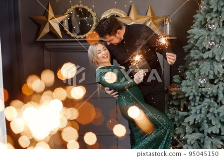 Young woman and man standing near christmas tree and holding a sparkler. Cheerful family celebrating Christmas. Blonde woman wearing fashion green dress and bearded man wearing black shirt. Young woman and man standing near christmas tree and holding a sparkler. Cheerful family celebrating Christmas. Blonde woman wearing fashion green dress and bearded man wearing black shirt. 95045150