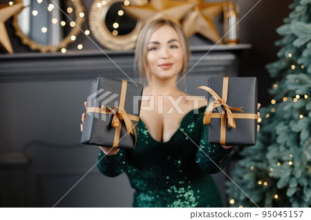 Portrait of woman holding a gift boxes. Gorgeous blonde girl in an elegant long green dress celebrates Christmas. Woman standing by a beautiful Christmas tree in expensive interiors. 95045157