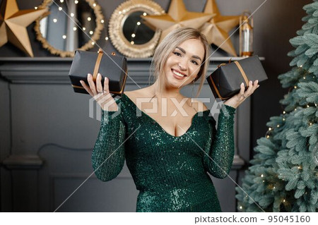 Portrait of woman holding a gift boxes. Gorgeous blonde girl in an elegant long green dress celebrates Christmas. Woman standing by a beautiful Christmas tree in expensive interiors. Portrait of woman holding a gift boxes. Gorgeous blonde girl in an elegant long green dress celebrates Christmas. Woman standing by a beautiful Christmas tree in expensive interiors. 95045160