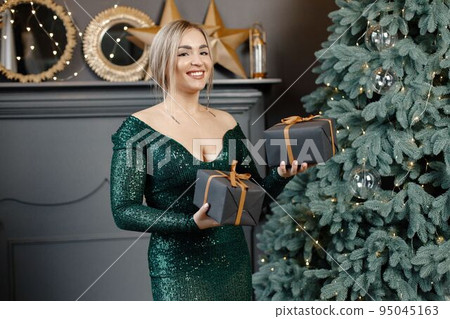 Portrait of woman holding a gift boxes. Gorgeous blonde girl in an elegant long green dress celebrates Christmas. Woman standing by a beautiful Christmas tree in expensive interiors. 95045163
