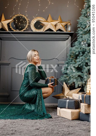 Portrait of woman holding a gift box. Gorgeous blonde girl in an elegant long green dress celebrates Christmas. Woman standing by a beautiful Christmas tree in expensive interiors. Portrait of woman holding a gift box. Gorgeous blonde girl in an elegant long green dress celebrates Christmas. Woman standing by a beautiful Christmas tree in expensive interiors. 95045166