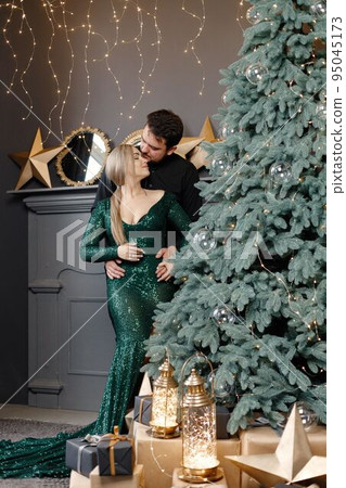 Young woman and man standing near christmas tree and kissing. Cheerful family celebrating Christmas. Blonde woman wearing fashion green dress and bearded man wearing black shirt. Young woman and man standing near christmas tree and kissing. Cheerful family celebrating Christmas. Blonde woman wearing fashion green dress and bearded man wearing black shirt. 95045173