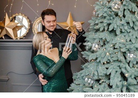Young woman and man standing near christmas tree and hugging. Cheerful family celebrating Christmas. Blonde woman wearing fashion green dress and bearded man wearing black shirt. 95045189