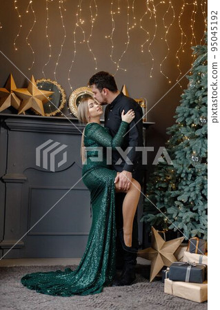 Young woman and man standing near christmas tree and hugging. Cheerful family celebrating Christmas. Blonde woman wearing fashion green dress and bearded man wearing black shirt. 95045192