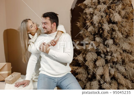 Young woman and man sitting near christmas tree and hugging. Cheerful family celebrating Christmas. Blonde woman and bearded man wearing white clothes. 95045235