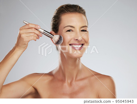 Face, makeup and brush with a mature woman applying blusher to her cheek in studio on a gray background. Beauty, skin and cosmetics with an attractive female posing with a product and a smile 95045943