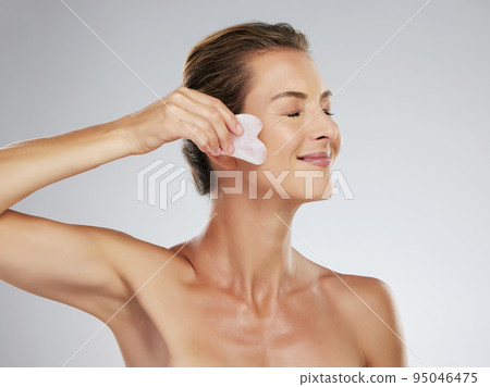 Beauty, face and massage with a woman using a gua sha or scraping tool in studio on a gray background for skincare. Wellness, skin and body with a mature female taking care in her antiaging routine Beauty, face and massage with a woman using a gua sha or scraping tool in studio on a gray background for skincare. Wellness, skin and body with a mature female taking care in her antiaging routine 95046475