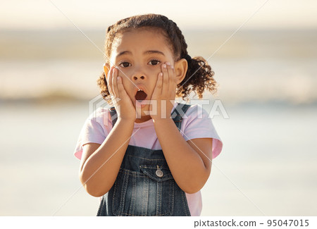 Wow, children and hands on the face of a girl on the beach looking surprised or shocked outside during summer. Kids, youth and surprise with a little female child feeling shock while outdoor alone 95047015