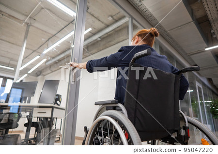 Red-haired caucasian woman in a wheelchair trying to open the door in the office. Red-haired caucasian woman in a wheelchair trying to open the door in the office. 95047220