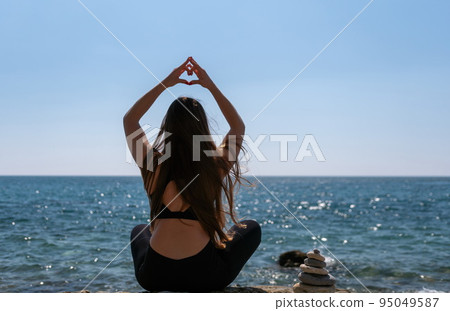 Woman makes heart with hands on beach. Young woman with long hair, fitness instructor, stretching before pilates, on a yoga mat near the sea on a sunny day, female fitness yoga routine concept Woman makes heart with hands on beach. Young woman with long hair, fitness instructor, stretching before pilates, on a yoga mat near the sea on a sunny day, female fitness yoga routine concept 95049587