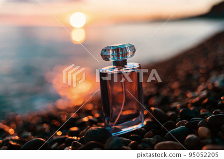 Presentation of the summer fragrance. A glass bottle of golden perfume on a pebble beach, against the sunset. The concept of selective perfumery and the fashionable luxury industry 95052205