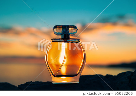 Rounded glass bottle of perfume on the ocean's beach. Sunset and sky in the background. Close-up low angle view. Presentation of perfumery 95052206