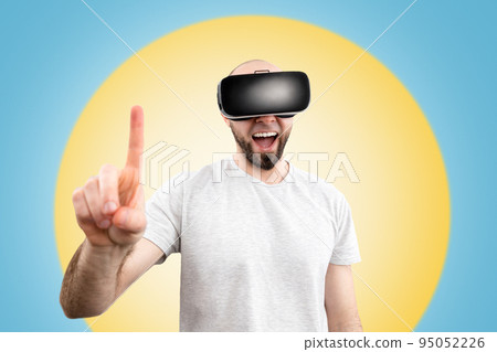 VR glasses. A wondered man in virtual reality glasses, with his mouth slightly open, points a finger into space. Blue background with a yellow circle. The concept of virtual reality 95052226