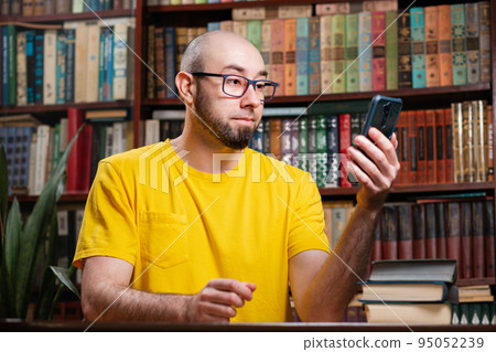 Communication. A bald bearded man with glasses uses a smartphone. In the background is a library and shelves of books. Concept of social networks, distance learning and quarantine 95052239
