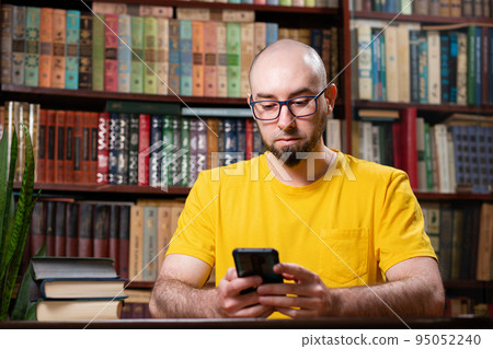 Communication. A bald man with glasses uses a smartphone. In the background is a library and shelves of books. Concept of social networks, distance learning and quarantine 95052240