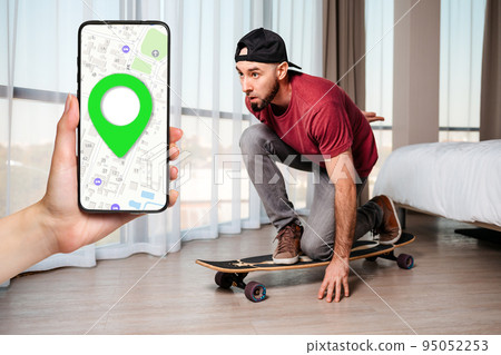 A young man sitting down posing on a longboard. Home interior. A female's hand holds a smartphone with an online map. Concept of quarantine and online navigation 95052253