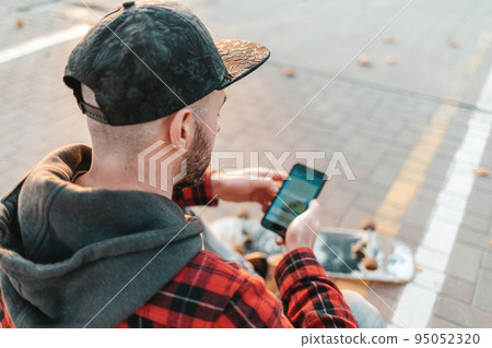 martphone and communication. A young guy in a cap, with a beard, uses a smartphone. In the background is a road and a skateboard. Back view from over the shoulder martphone and communication. A young guy in a cap, with a beard, uses a smartphone. In the background is a road and a skateboard. Back view from over the shoulder 95052320