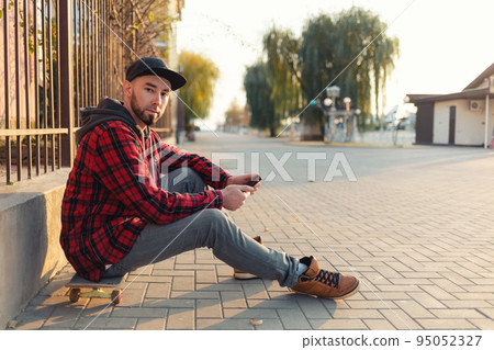 Skateboarding. A young man sits on a skateboard, with a smartphone in his hands. The man is wearing a black cap, jeans and a red checked shirt. Outdoor 95052327