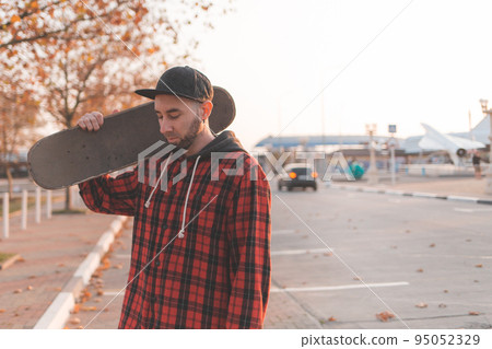 Skateboarding. A handsome guy with a beard, in a cap and fashionable clothes posing with a skateboard in his hands. In the background, an empty street of the city. Copy space Skateboarding. A handsome guy with a beard, in a cap and fashionable clothes posing with a skateboard in his hands. In the background, an empty street of the city. Copy space 95052329