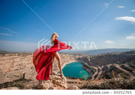 Side view of a beautiful sensual woman in a red long dress posing on a rock high above the lake in the afternoon. Against the background of the blue sky and the lake in the form of a heart Side view of a beautiful sensual woman in a red long dress posing on a rock high above the lake in the afternoon. Against the background of the blue sky and the lake in the form of a heart 95052765