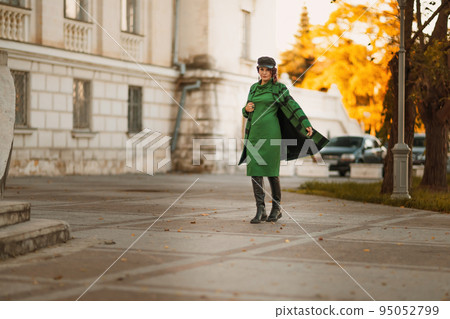 Outdoor fashion portrait of an elegant fashionable brunette woman, model in a stylish cap, green dress, posing at sunset in a European city in autumn. 95052799