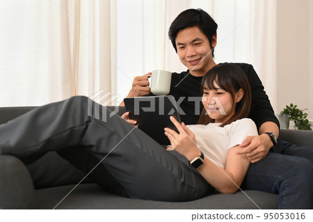 Loving asian couple embracing and browsing internet on digital tablet in bright living room. People and technology concept 95053016