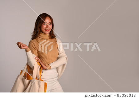 Portrait of attractive young woman wearing warm clothes holding white tote bag standing over grey background 95053018