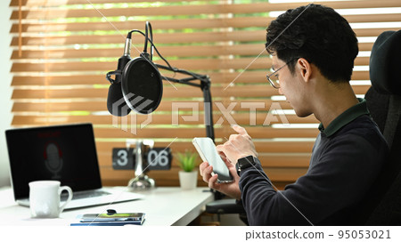 Side view of man radio host sitting front of condenser microphone and using smart phone Side view of man radio host sitting front of condenser microphone and using smart phone 95053021