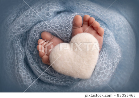 The tiny foot of a newborn baby. Soft feet of a new born in a blue wool blanket. Close up of toes, heels and feet of a newborn. Knitted white heart in the legs of a baby. Macro photography. The tiny foot of a newborn baby. Soft feet of a new born in a blue wool blanket. Close up of toes, heels and feet of a newborn. Knitted white heart in the legs of a baby. Macro photography. 95053465