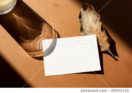 Still life scene with hard shadows on brown background in sunlight glass and blank business, greeting card, invitation mockup. Long harsh shadows. New naturalism Still life scene with hard shadows on brown background in sunlight glass and blank business, greeting card, invitation mockup. Long harsh shadows. New naturalism 95055120
