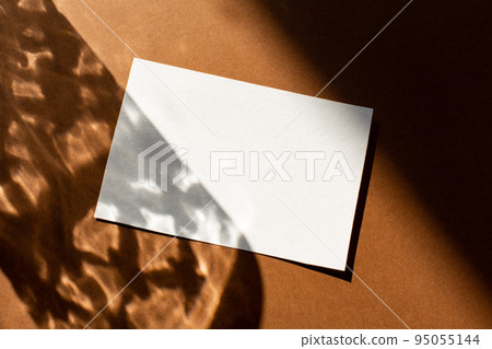 Still life scene with hard shadows on brown background in sunlight glass and blank business, greeting card, invitation mockup. Long harsh shadows. New naturalism  95055144