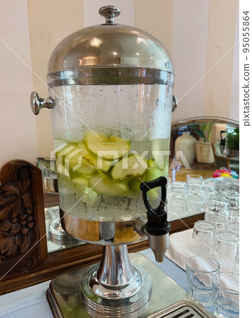 A glass transparent tank with a tap with cold drinking water with pieces of fruit and mint stands in the hotel lobby. A glass transparent tank with a tap with cold drinking water with pieces of fruit and mint stands in the hotel lobby. 95055864