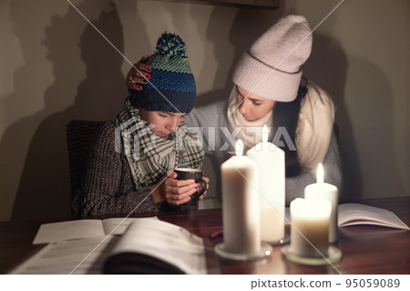 Worried mother comforts her freezing son holding tea and studying under candlelights in the war zone, both wearing winter clothes at home 95059089