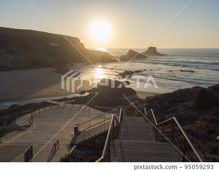 Sunset view of sand beach Praia da Zambujeira do Mar with rock and cliff, stairs and the setting sun to the ocean waves. Rota Vicentina coast, Odemira, Portugal Sunset view of sand beach Praia da Zambujeira do Mar with rock and cliff, stairs and the setting sun to the ocean waves. Rota Vicentina coast, Odemira, Portugal 95059293