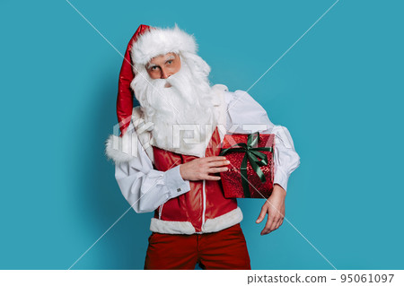 Christmas. Stylish santa claus holds red gift box on color background while looking at camera. 95061097