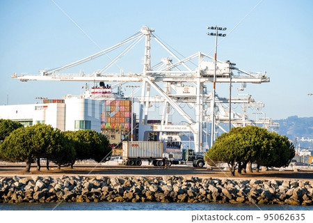 San Francisco, USA. Loaded trucks moving by Container cranes at bay area. View of busy Port of Oakland in city with blue sky in background. Shipping terminal facility on sunny day. 95062635