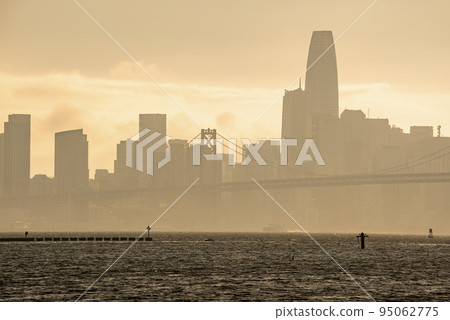 Bay Bridge and downtown district on coastal seafront. Famous tourist attraction connecting urban San Francisco. Scenic view of city skyline with sky in background during sunset. 95062775
