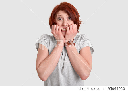 Photo of frightened red haired woman bites fingers nails, looks with nervous expression, afraids of difficulties, dressed in casual t shirt, models over white background. Negative feelings concept Photo of frightened red haired woman bites fingers nails, looks with nervous expression, afraids of difficulties, dressed in casual t shirt, models over white background. Negative feelings concept 95063030