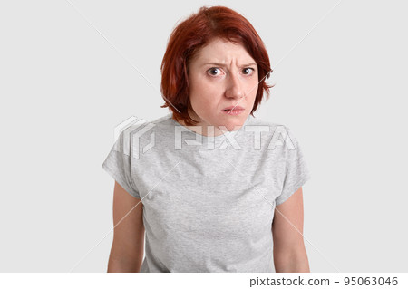 Photo of beautiful redhead woman looks with curious gaze, bewilderment at camera, wears casual t shirt, models over white studio background, has displeased facial expression. People and emotions Photo of beautiful redhead woman looks with curious gaze, bewilderment at camera, wears casual t shirt, models over white studio background, has displeased facial expression. People and emotions 95063046