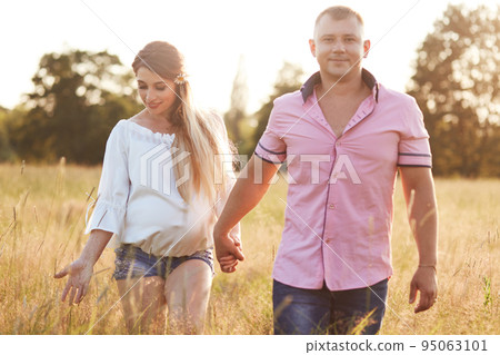 Happy young future parents have stroll across green meadow, hold hands, enjoy calm atmosphere, have happy expressions, anticipate for baby. People, family, relationship and pregnancy concept Happy young future parents have stroll across green meadow, hold hands, enjoy calm atmosphere, have happy expressions, anticipate for baby. People, family, relationship and pregnancy concept 95063101