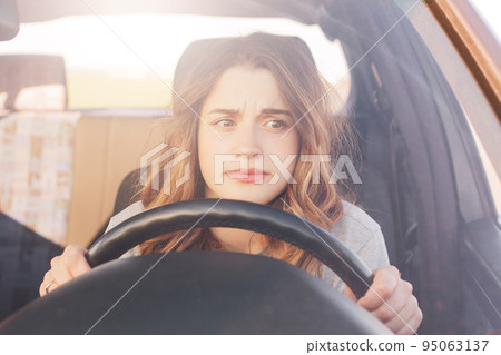 Nervous female driver sits at wheel, has worried expression as afraids to drive car by herself for first time. Frightened woman has car accident on road. People, driving, problems with transport Nervous female driver sits at wheel, has worried expression as afraids to drive car by herself for first time. Frightened woman has car accident on road. People, driving, problems with transport 95063137