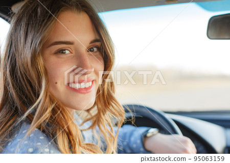 Back view of attractive woman in car, sitting at wheel, turning and looking at passengers in back seat. Beautiful female looks at camera with toothy smile, being glad to pass exam in driving school. Back view of attractive woman in car, sitting at wheel, turning and looking at passengers in back seat. Beautiful female looks at camera with toothy smile, being glad to pass exam in driving school. 95063159