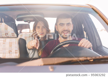 Astonished young woman sits on back seat in car, indicates at windscreen with interested expression as sees big animal on road and her unshaven husband in foreground drives car, has surprised look. 95063292