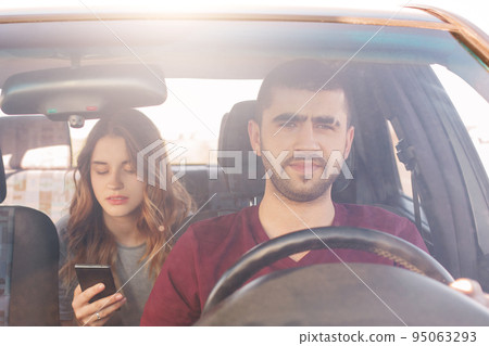 Front view of focused male driver sits at wheel and drives car while his girlfriend sits on back seat, holds smart phone and messages with friends or finds way in online map. Enjoy travel together 95063293