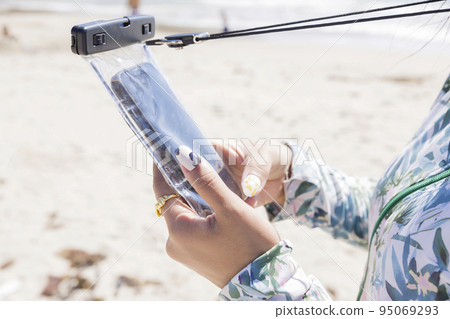 A woman's hand looking at a smartphone in a waterproof case on the beach 95069293