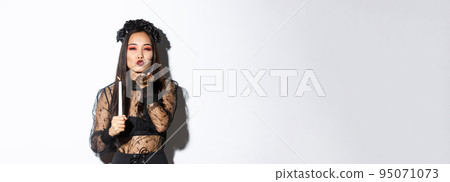 Beautiful and elegant asian woman in gothic lace dress celebrating halloween, dress-up as witch, sending air kiss at camera and holding candle 95071073