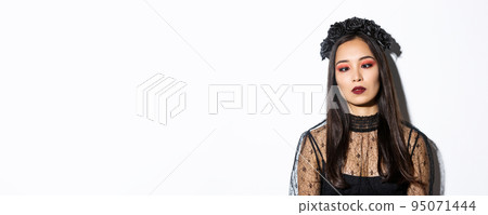 Close-up of bored and annoyed asian woman in halloween costume of witch squinting, trying to look at nose, standing over white background 95071444
