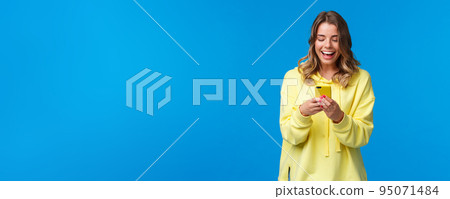 Technology and people concept. Happy smiling blond girl using mobile phone in yellow case, messaging look at smartphone display with pleased expression, send funny meme, blue background Technology and people concept. Happy smiling blond girl using mobile phone in yellow case, messaging look at smartphone display with pleased expression, send funny meme, blue background 95071484